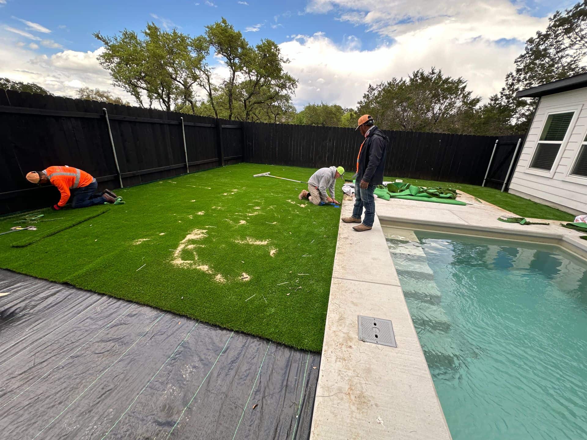 Workers installing green artificial turf beside a backyard pool and patio under a cloudy sky
