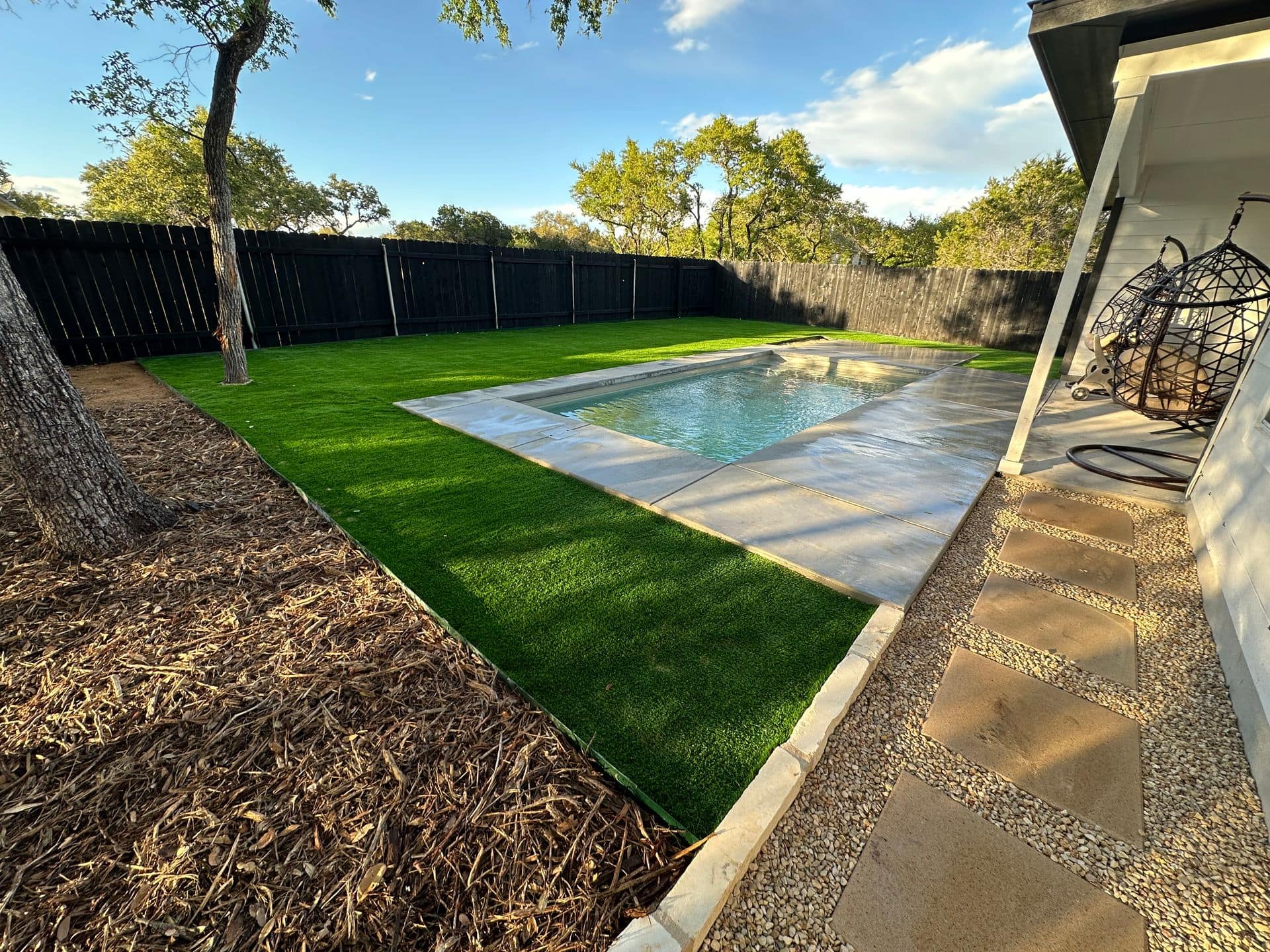 Backyard with pool and green lawn, bordered by a black fence and trees.