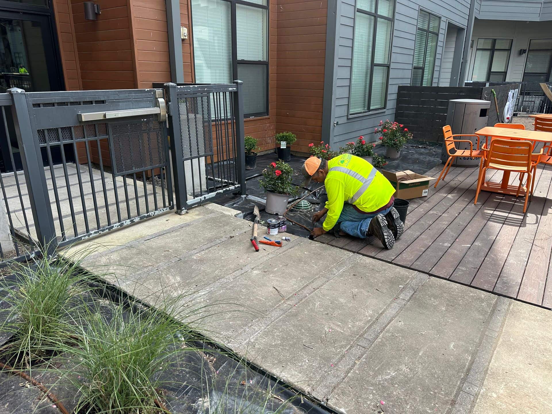 Person in neon vest works near a fenced patio, next to a building with orange tables.