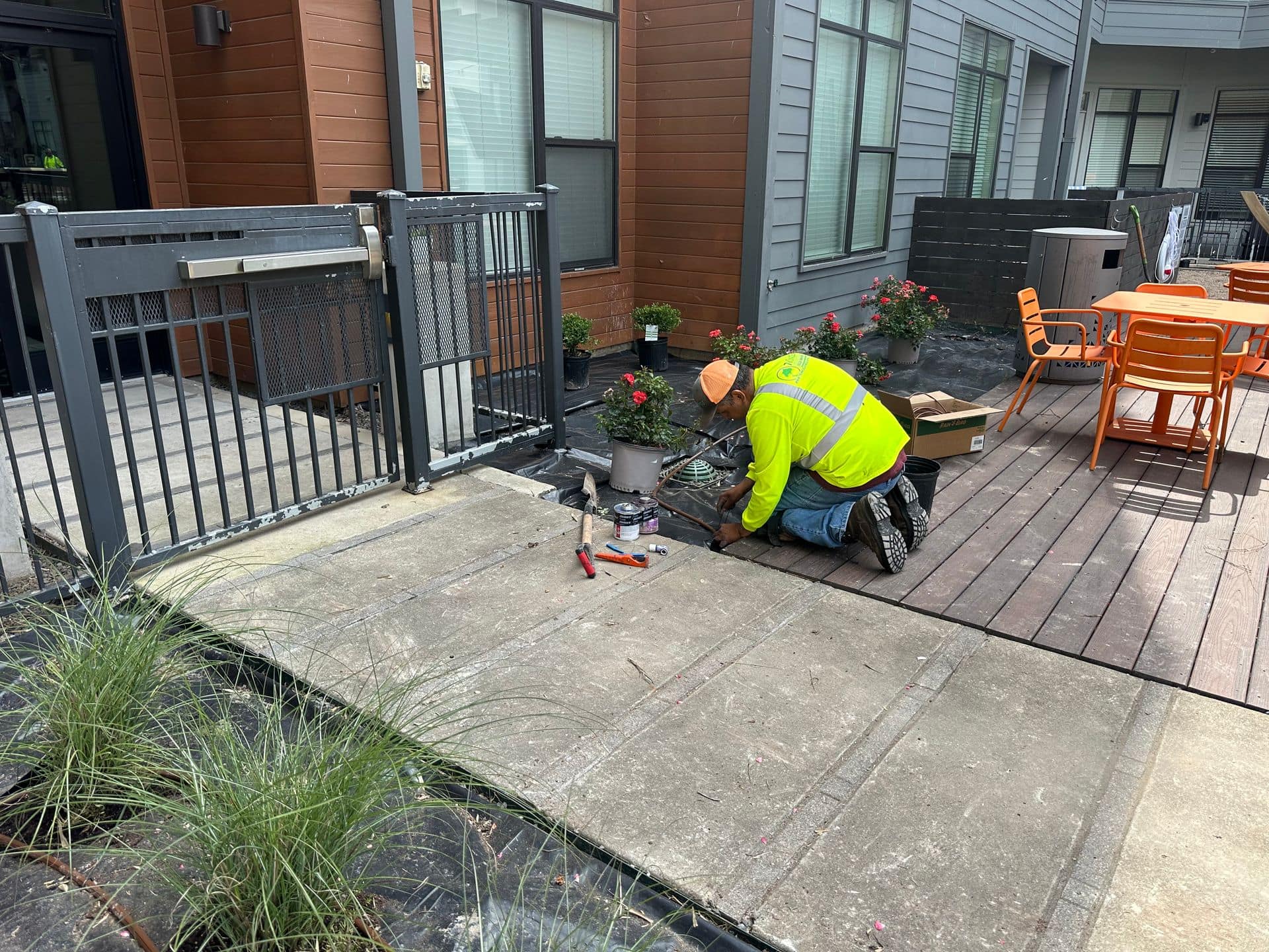 Person in neon vest works near a fenced patio, next to a building with orange tables.